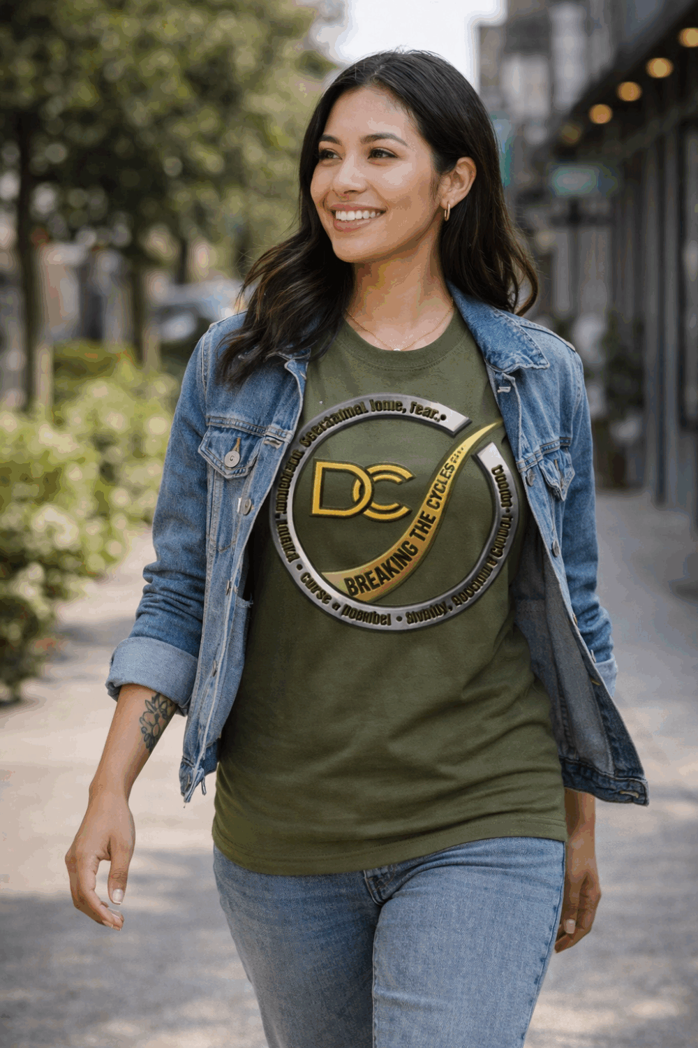Latina woman walking outdoors wearing olive green Breaking The Cycles DCC t-shirt with denim jacket